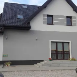 Dwukondygnacyjny dom w Łęczycy z szarą elewacją, ciemną dachówką i oryginalnymi poziomymi zdobieniami okiennymi.