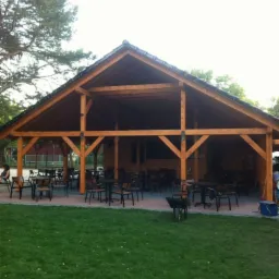 Drewniana konstrukcja z zadaszeniem z ciemnej dachówki, tworząca otwartą przestrzeń z licznymi stolikami i krzesłami, otoczona trawnikiem i drzewami.