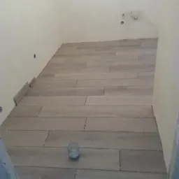 Układanie podłogi z szarych, drewnopodobnych płytek w małej łazience, widoczne krzyżyki dystansowe i resztki zaprawy, szklanka z brudną wodą na środku podłogi.