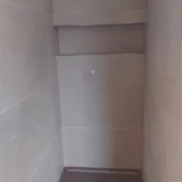 Wnęka prysznicowa z odpływem liniowym w podłodze, wykończona szarymi płytkami ceramicznymi na ścianach i ciemniejszą podłogą, widoczne dwa otwory na armaturę.