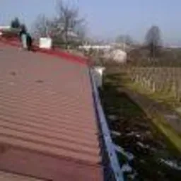 Czerwony dach z blachy trapezowej z nowymi, białymi rynnami i orynnowaniem, widok z góry na tle drzew i błękitnego nieba; w tle widoczna postać pracownika.