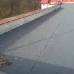 Czarny, płaski dach pokryty papą termozgrzewalną z widocznymi liniami łączeń, fragment czerwonej obróbki blacharskiej przy krawędzi dachu, liście na powierzchni.