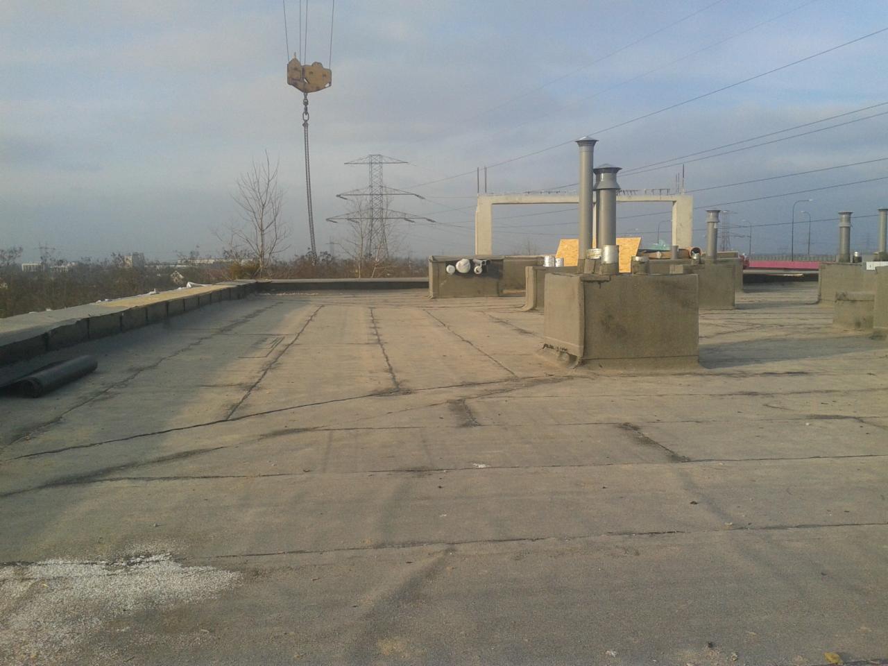 Płaski dach pokryty papą z kominami wentylacyjnymi, w tle słup wysokiego napięcia i żuraw budowlany.