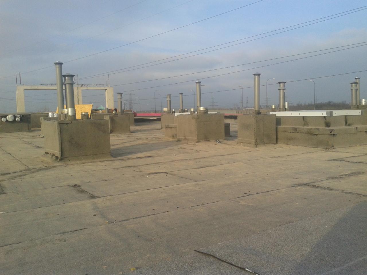 Płaski dach budynku przemysłowego z betonowymi podstawami pod kominy wentylacyjne i rurami, widoczne elementy konstrukcyjne i instalacyjne, w tle słupy wysokiego napięcia.