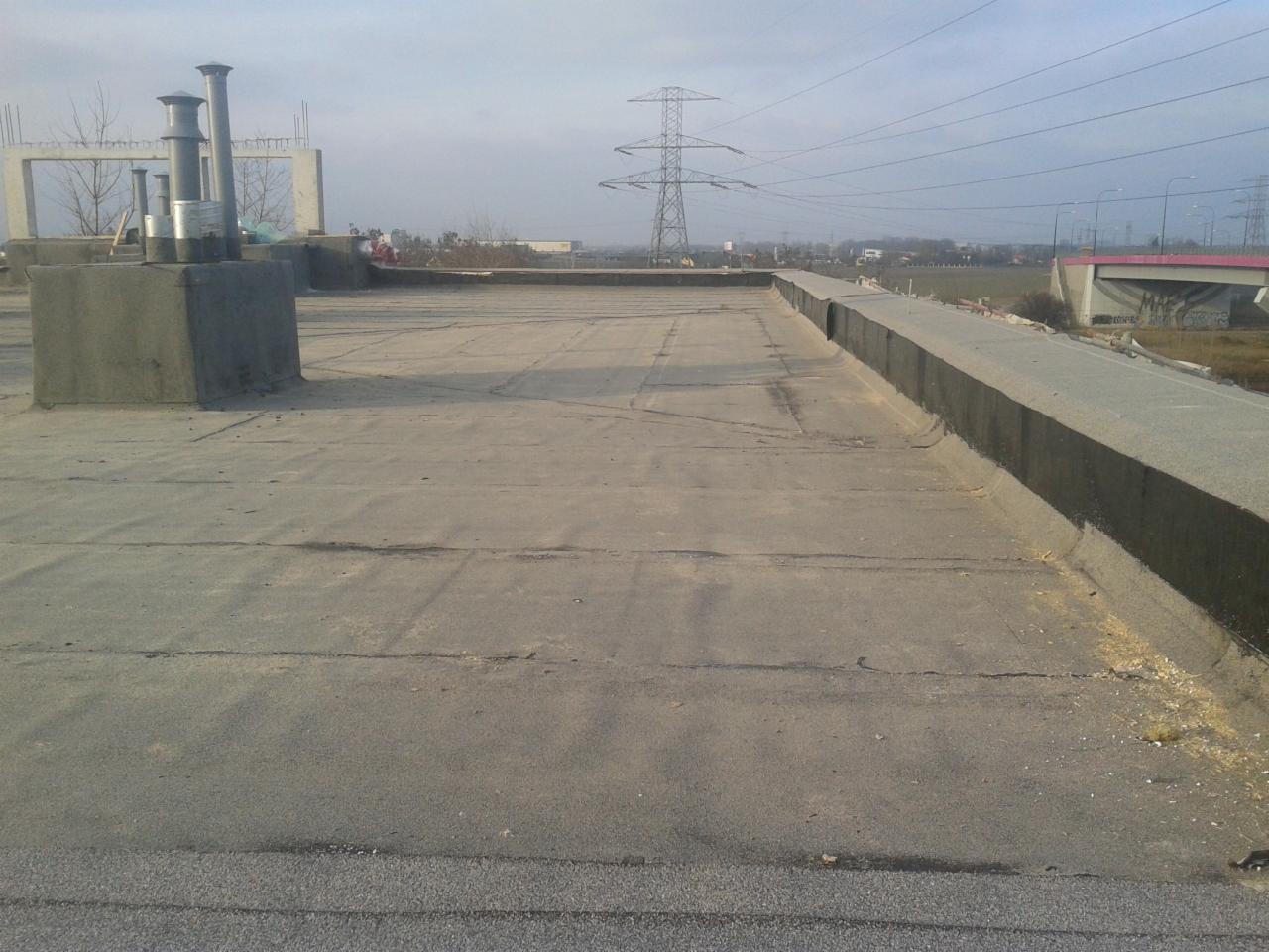 Płaski dach pokryty papą z widocznymi kominami wentylacyjnymi i betonową attyką, w tle słupy wysokiego napięcia i wiadukt.