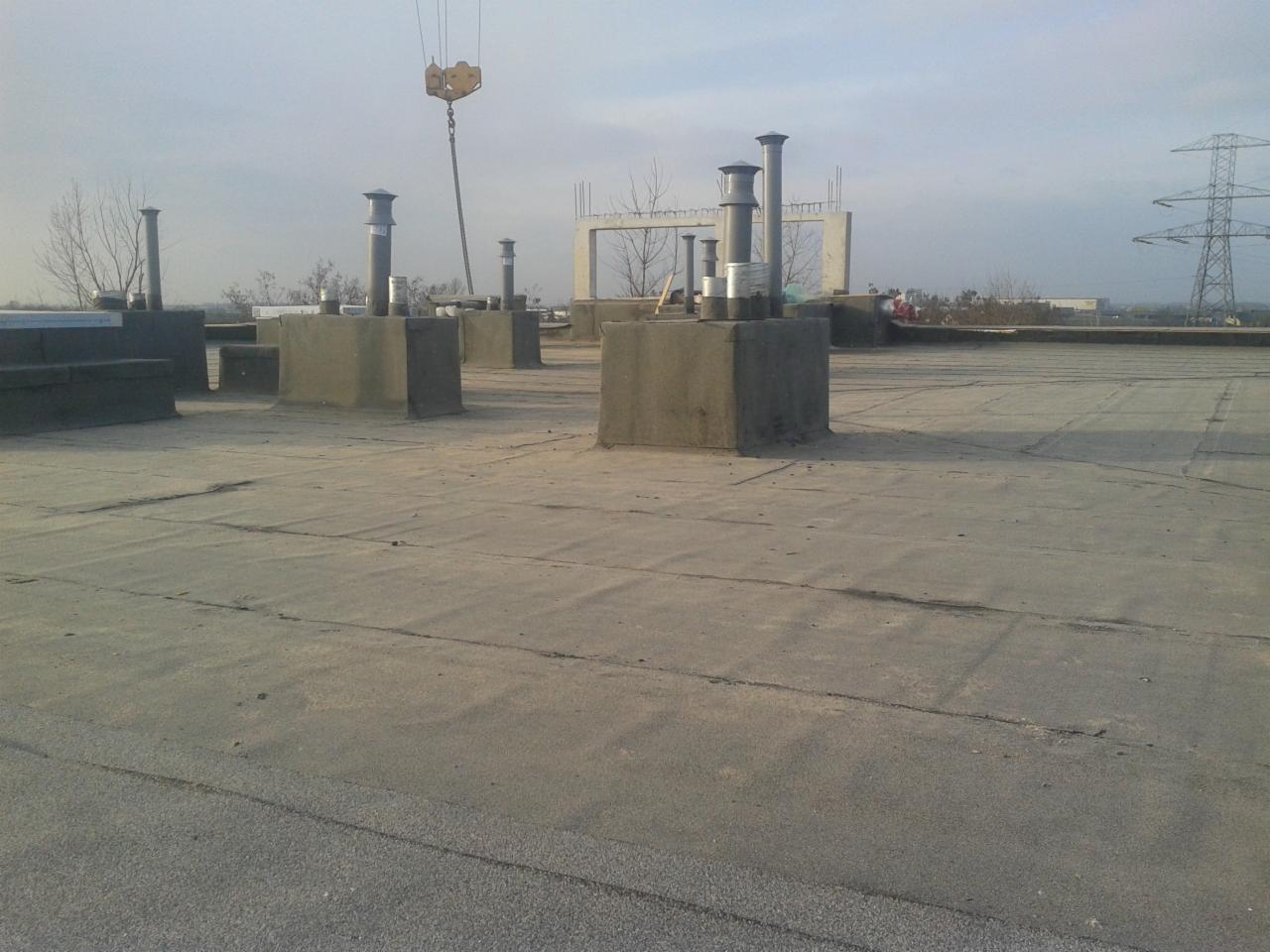 Płaski dach pokryty papą z wystającymi kominami wentylacyjnymi i elementami konstrukcyjnymi w tle, widoczny dźwig budowlany w trakcie pracy.