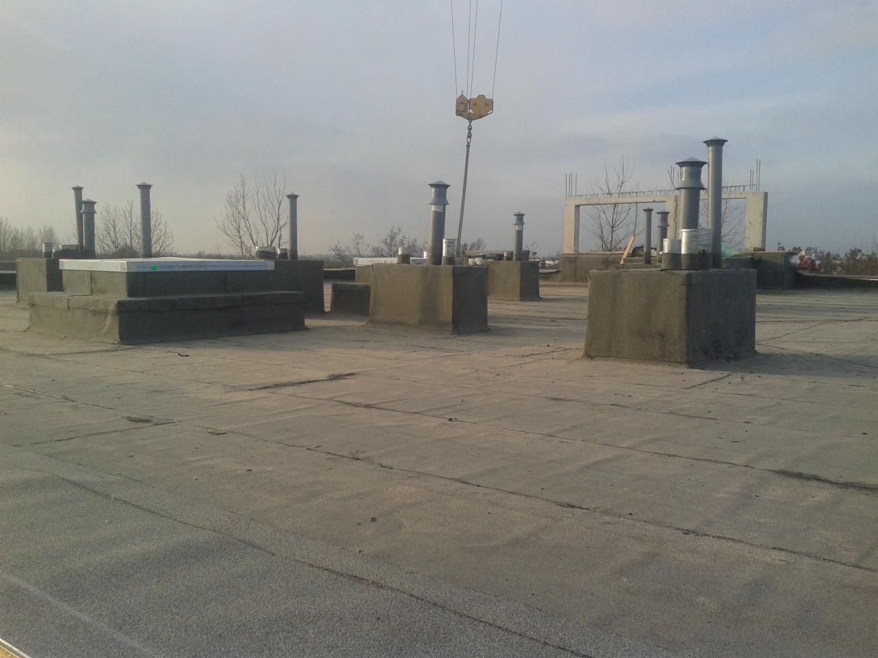 Płaski dach pokryty papą z licznymi kominami wentylacyjnymi i jednym świetlikiem, widoczny dźwig budowlany w tle, pochmurny dzień.