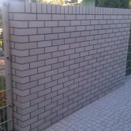 Klinkier bojs - Nowoczesne ogrodzenie z szarej cegły i metalowej siatki, widok z bliska na połączenie materiałów i fakturę brukowanej nawierzchni.