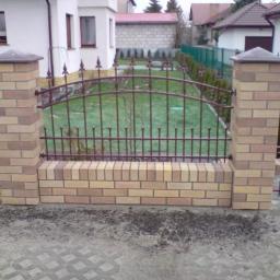 Klinkier bojs - Fragment ogrodzenia z ceglanymi słupkami i metalowymi przęsłami na tle zielonego trawnika i zabudowań mieszkalnych.
