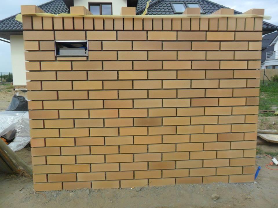 Wznoszona z cegły klinkierowej ściana ogrodzenia z otworem na skrzynkę pocztową, widoczna na tle domu z ciemnym dachem i fragmentem terenu budowy.