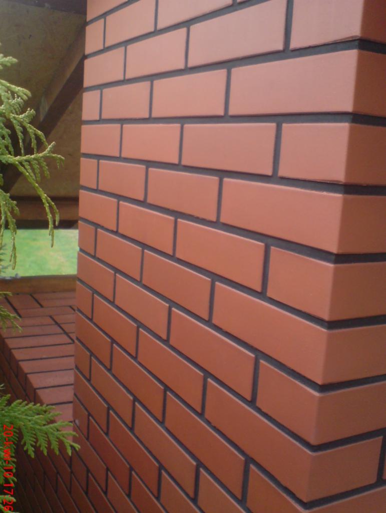Fragment ogrodzenia z klinkieru w odcieniach ceglanej czerwieni, z ciemną fugą, widziany pod kątem. W tle zieleń i fragment konstrukcji.