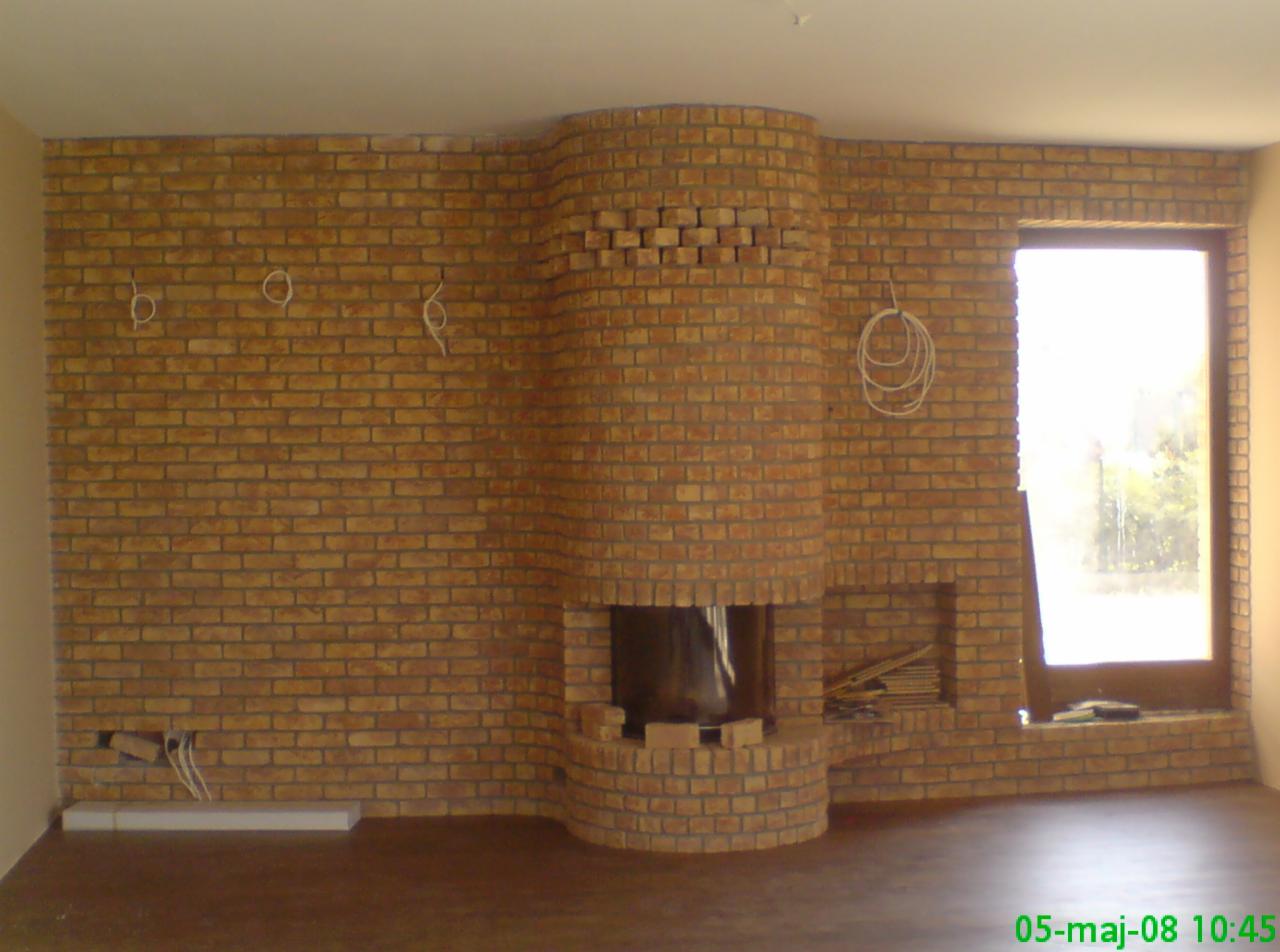 Murowany kominek z cegły klinkierowej w trakcie budowy; widoczne przewody elektryczne, okno z widokiem na zieleń i fragment podłogi. Prace murarskie.