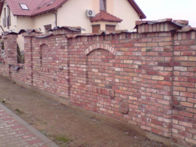 Murowane ogrodzenie z cegły klinkierowej z dekoracyjnymi łukami i filarami, widoczne z bliska na tle domu z czerwonym dachem.