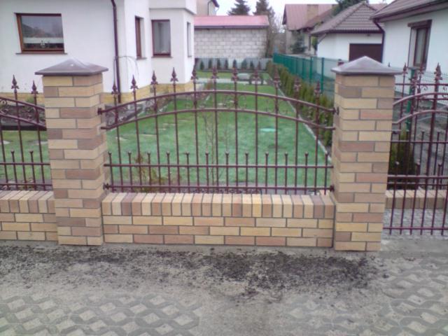 Fragment ogrodzenia z ceglanymi słupkami i metalowymi przęsłami na tle zielonego trawnika i zabudowań mieszkalnych.