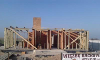 Wznoszenie szkieletu domu z drewnianą więźbą dachową i kominem z cegły, widoczne elementy konstrukcyjne i tablica informacyjna.