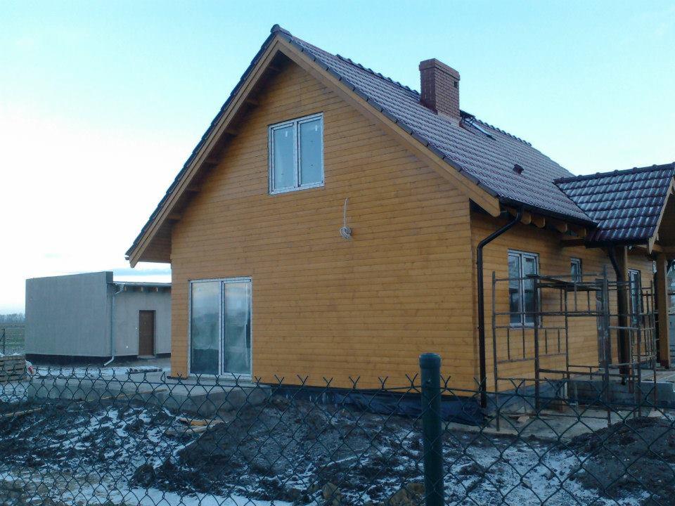 Surowy stan domu szkieletowego z drewnianą elewacją i częściowo ukończonym dachem z kominem, widoczne rusztowania przy bocznej ścianie, teren wokół ogrodzony siatką.