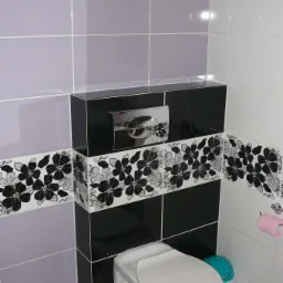 Wykończona łazienka: podwieszana toaleta z czarno-białą obudową w kwiaty, fioletowe i białe płytki na ścianach, wentylator w górnym rogu.