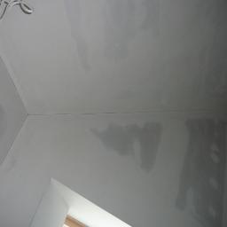 LUKSUS Styl  ŁUKASZ KORNILAK - Wnętrze po szpachlowaniu: gładkie, szare ściany i sufit z widocznymi przewodami. Ujęcie z dołu, fragment okna w rogu, przygotowane do malowania.