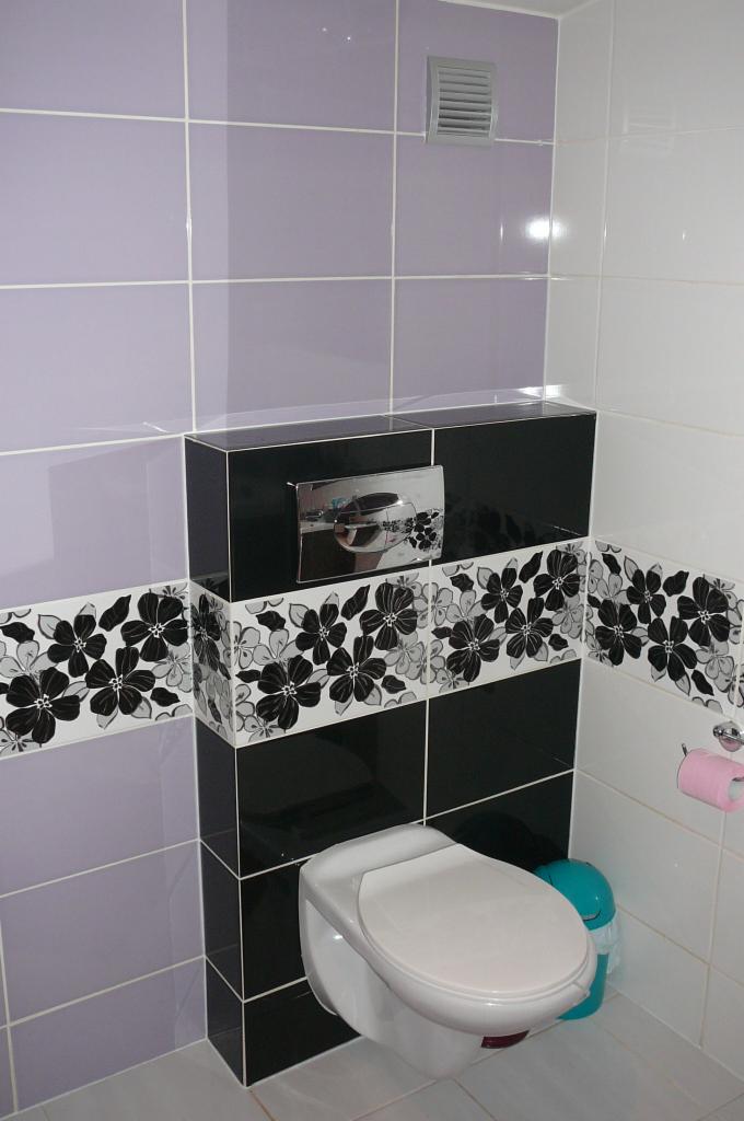 Wykończona łazienka: podwieszana toaleta z czarno-białą obudową w kwiaty, fioletowe i białe płytki na ścianach, wentylator w górnym rogu.