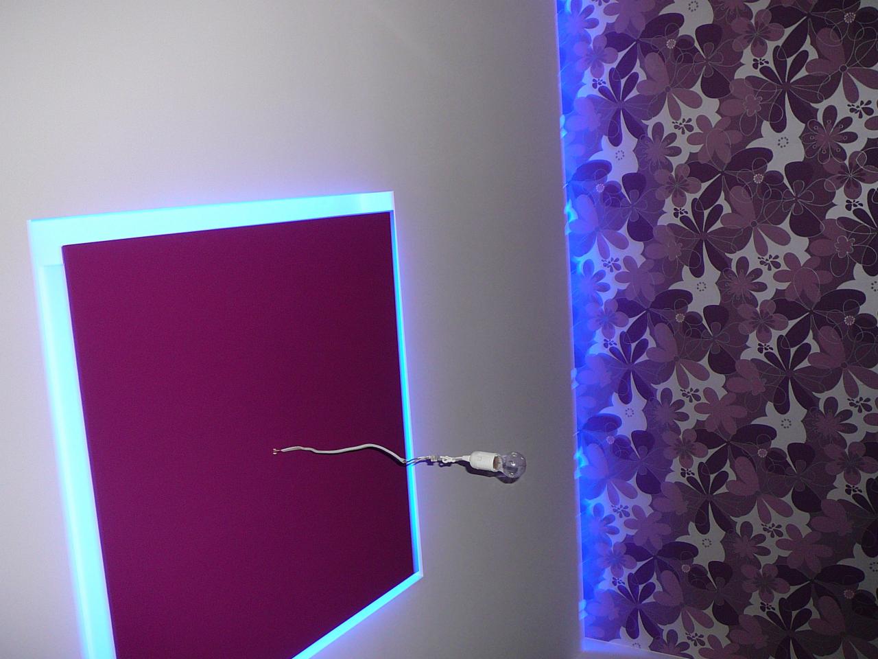 Fioletowy panel ścienny z niebieskim podświetleniem LED, obok ściana w kwiaty z podobnym podświetleniem. Widoczny przewód z żarówką.