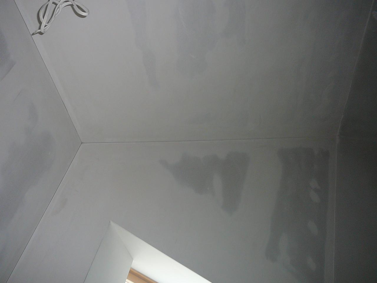 Wnętrze po szpachlowaniu: gładkie, szare ściany i sufit z widocznymi przewodami. Ujęcie z dołu, fragment okna w rogu, przygotowane do malowania.