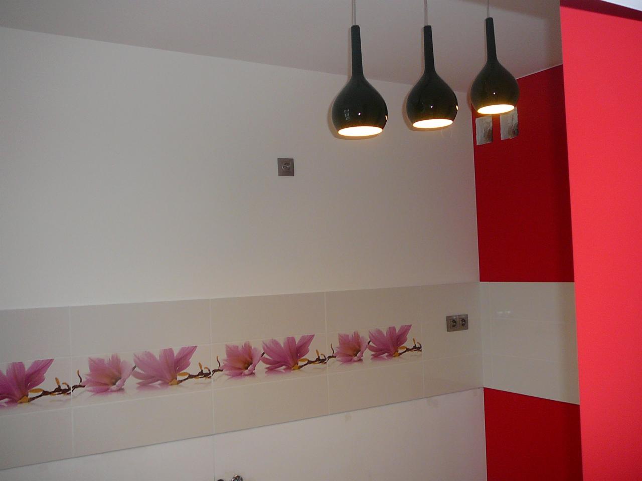 Wykończona łazienka z białymi płytkami zdobionymi motywem kwiatowym, czarnymi lampami wiszącymi i fragmentem czerwonej ściany. Widoczne gniazdka elektryczne.