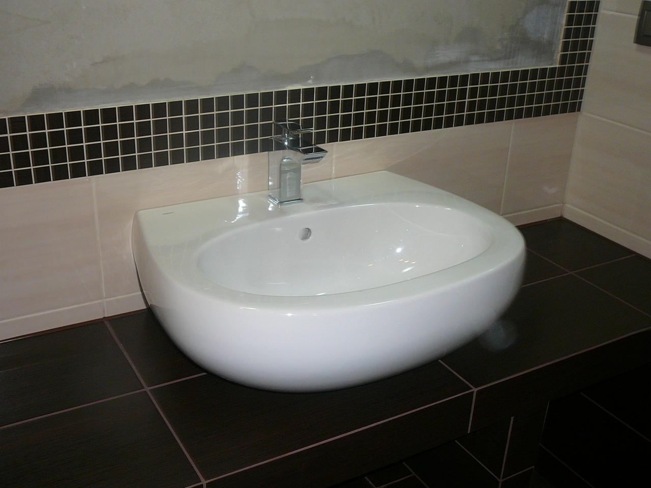 Nowoczesna, biała umywalka z chromowaną baterią na tle mozaiki i beżowych płytek w łazience. Widoczne ciemne płytki podłogowe.