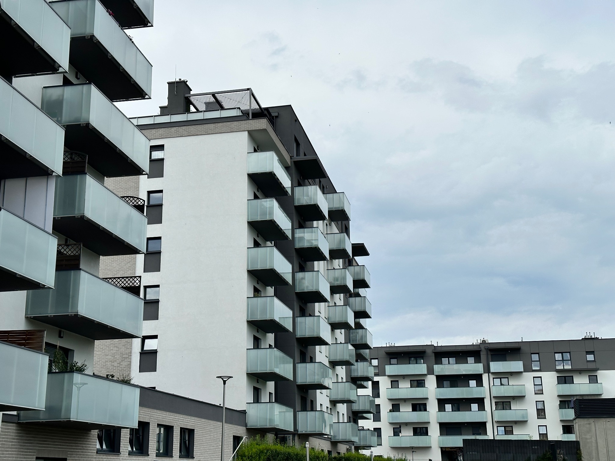 Nowoczesny budynek wielorodzinny z białymi elewacjami i balkonami z mlecznego szkła, widok od dołu na tle pochmurnego nieba.