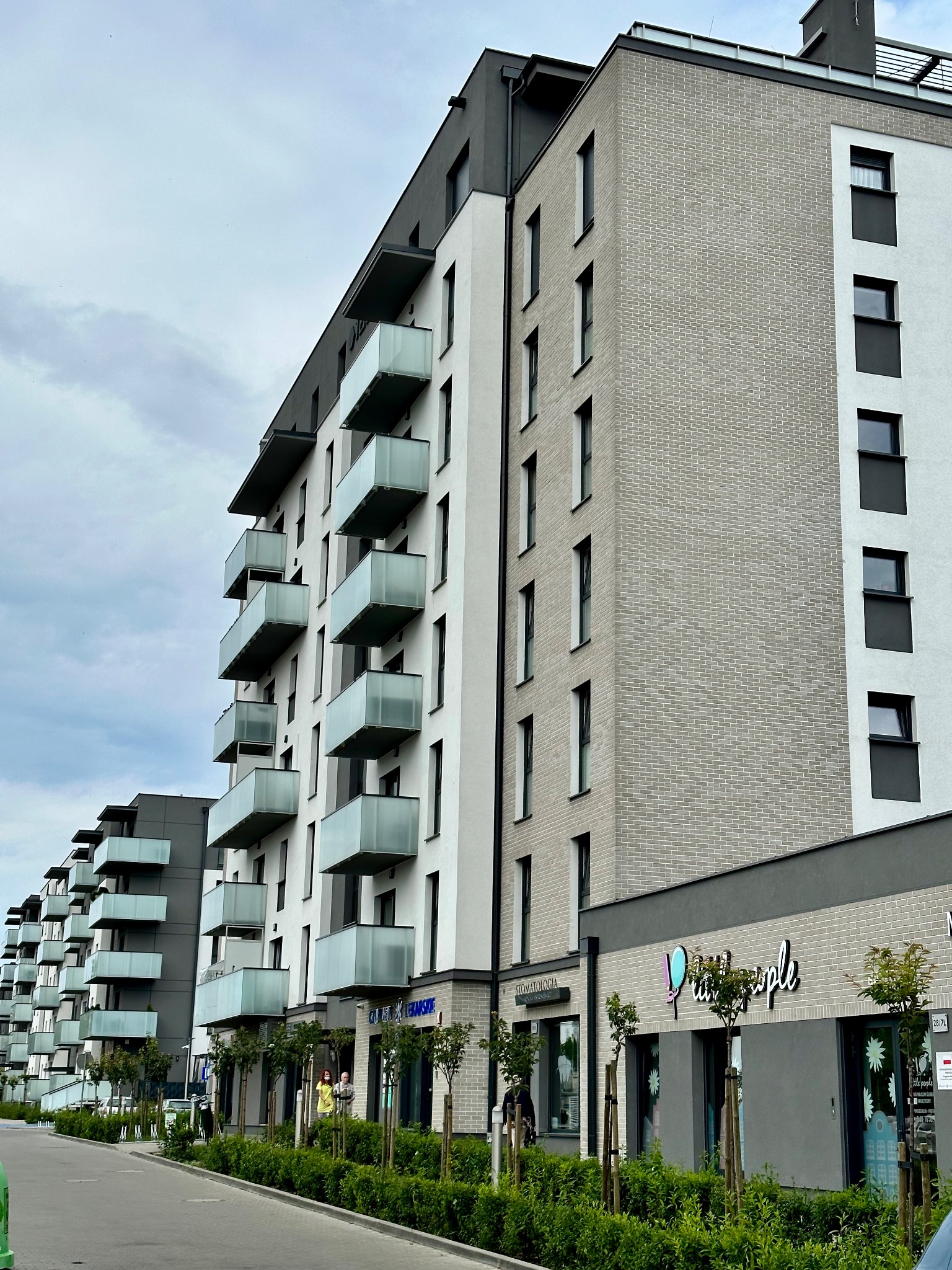 Nowoczesny budynek wielorodzinny z elewacją z szarej cegły i białego tynku, balkony z mlecznego szkła, widok z poziomu ulicy.