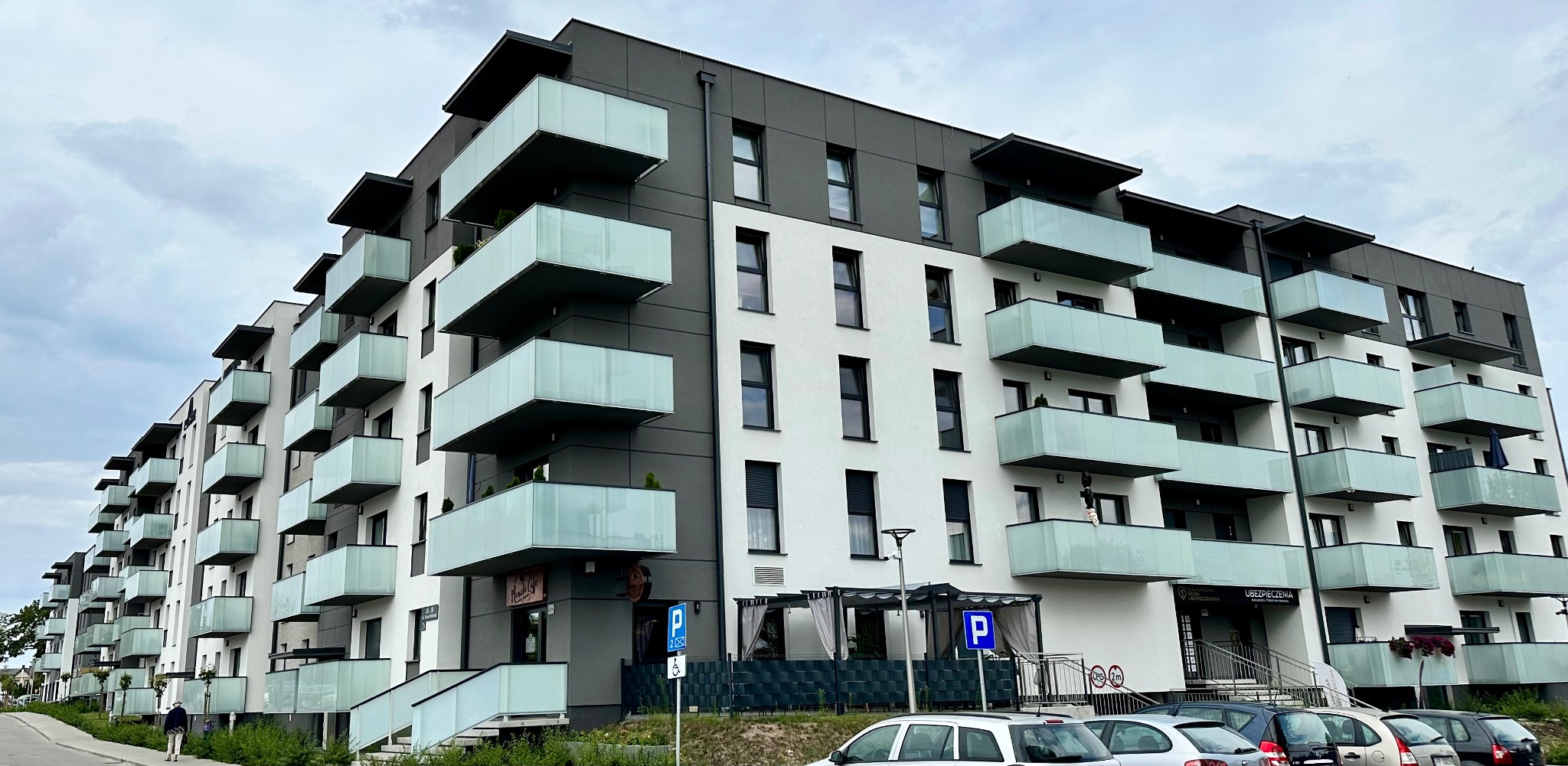 Nowoczesny, wielopiętrowy budynek mieszkalny z balkonami wykończonymi szkłem, białą elewacją i szarymi elementami architektonicznymi, widoczny na tle pochmurnego nieba.