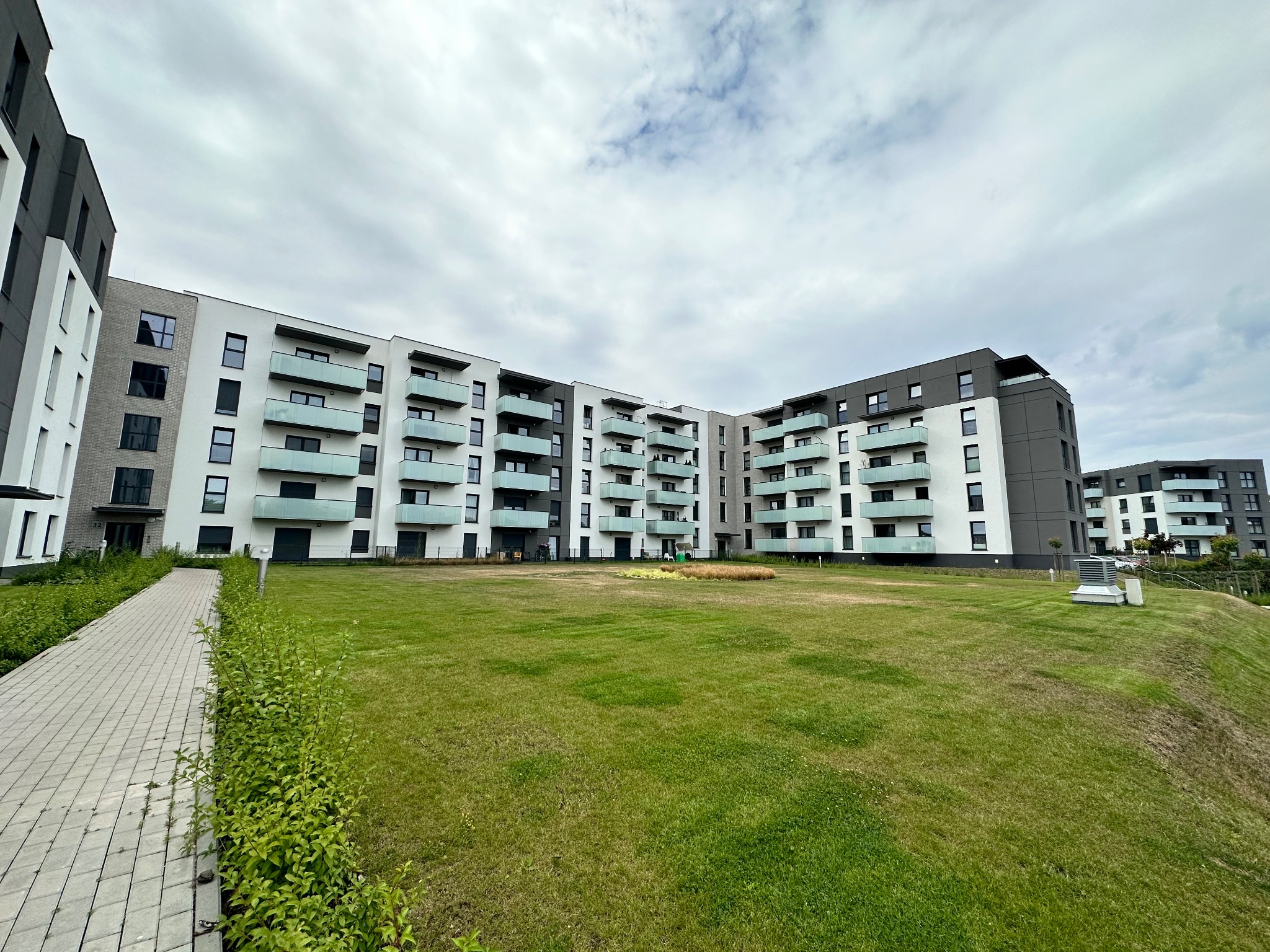 Nowoczesny budynek wielorodzinny z jasną elewacją, balkonami z przezroczystymi balustradami i szarymi akcentami, widok z poziomu trawnika w pochmurny dzień.