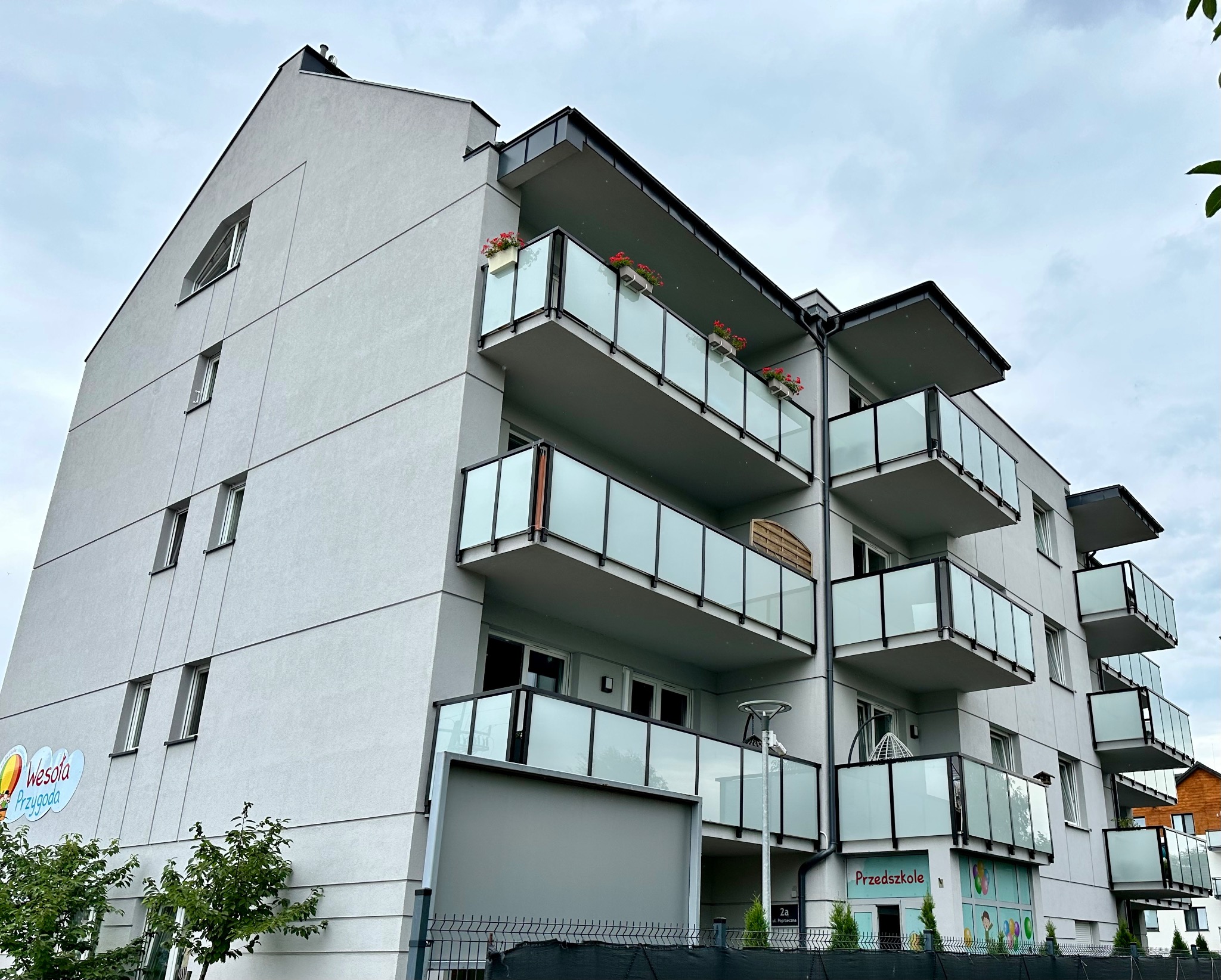 Nowoczesny budynek przedszkola z szarą elewacją, balkonami z przeszklonymi balustradami i kwiatami w donicach, widok z dołu na pochmurnym tle.