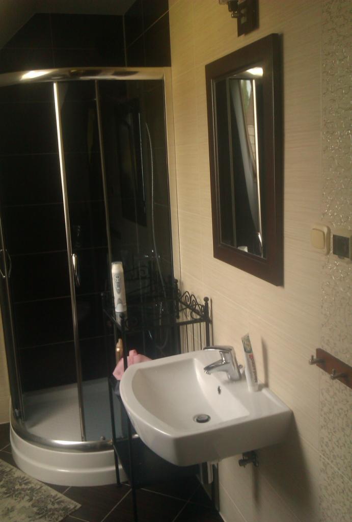 Nowoczesna łazienka z kabiną prysznicową, umywalką i lustrem w ciemnej ramie. Widoczne kremowe płytki ścienne i ciemne płytki podłogowe, tworzące kontrastowy design.