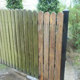 Częściowo wyczyszczone drewniane ogrodzenie, po lewej pokryte zielonym nalotem, po prawej odsłonięte naturalne drewno. Widoczne ślady mycia pod ciśnieniem.