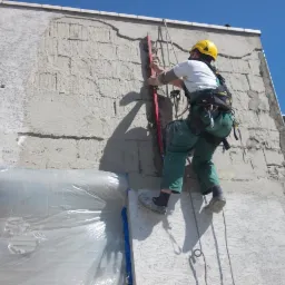 Pracownik w uprzęży, w kasku, z poziomicą, wyrównuje świeżo nałożony tynk na elewacji budynku. Część ściany zabezpieczona folią.