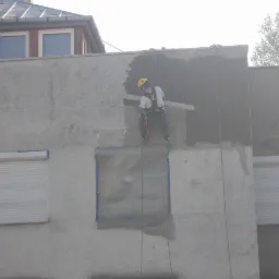 Pracownik w uprzęży, na linie, tynkuje elewację budynku. Okna zabezpieczone folią, widoczne fragmenty otynkowanej ściany. Praca na wysokościach.