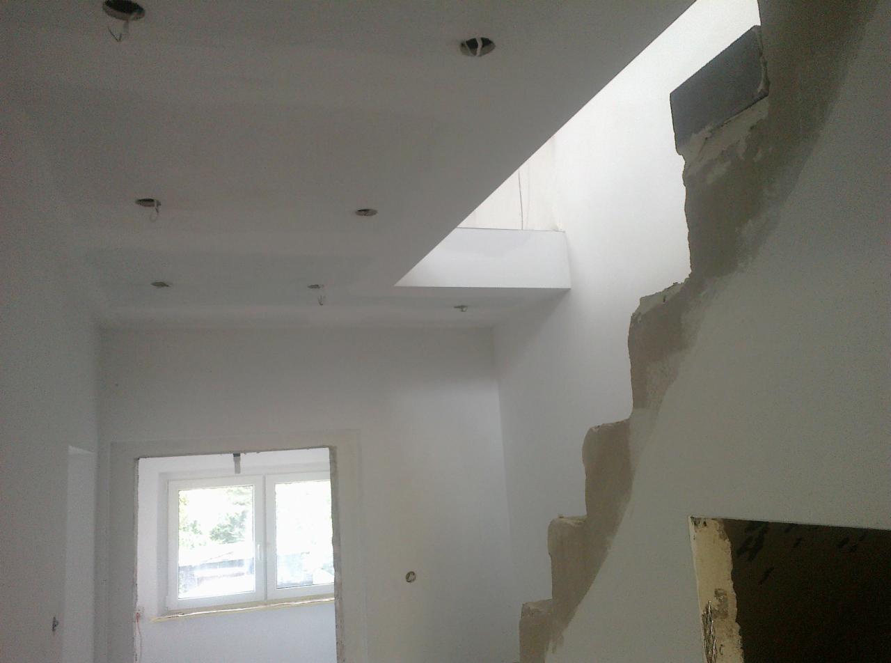 Wnętrze w trakcie prac wykończeniowych: białe ściany po szpachlowaniu, sufit z otworami na oświetlenie, fragment schodów i okno z widokiem na zieleń.