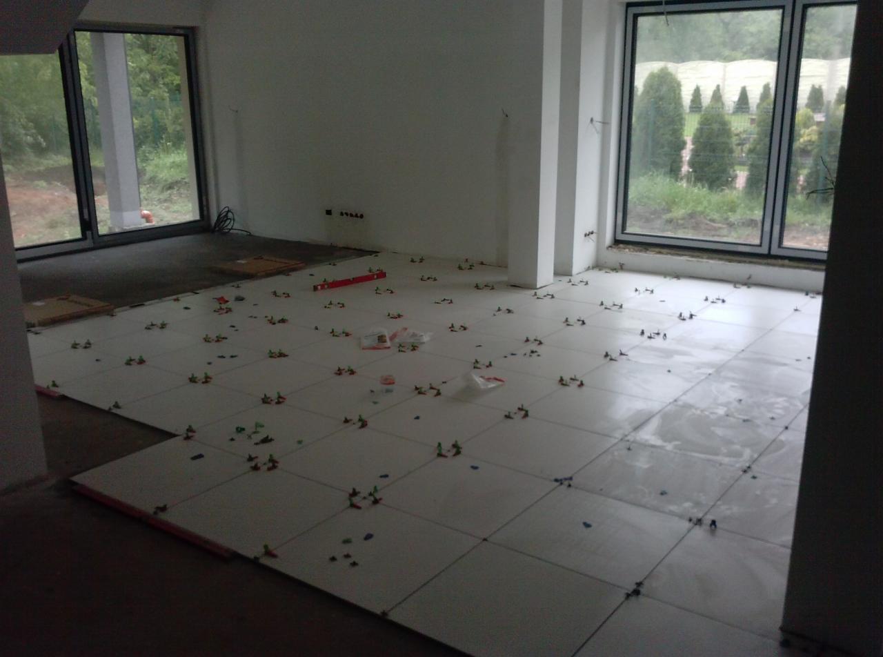 Układanie białych płytek podłogowych w jasnym pokoju z dużymi oknami. Widoczne krzyżyki dystansowe i poziomica. Część podłogi jeszcze nie wyłożona.