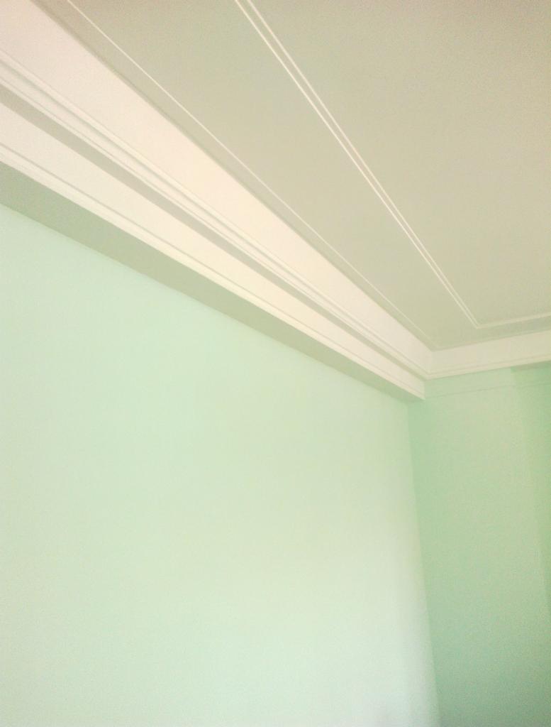 Pomalowany na jasnozielono róg pokoju z dekoracyjnym, białym wykończeniem sufitu. Widoczne listwy sufitowe i gładkie ściany.