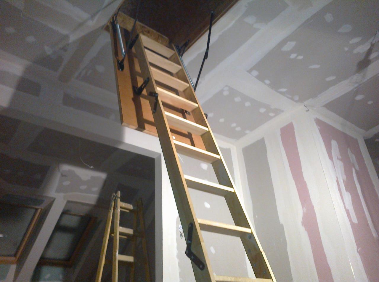 Drewniane schody strychowe rozłożone w pomieszczeniu w trakcie wykańczania, ściany i sufit z płyt kartonowo-gipsowych przed malowaniem.