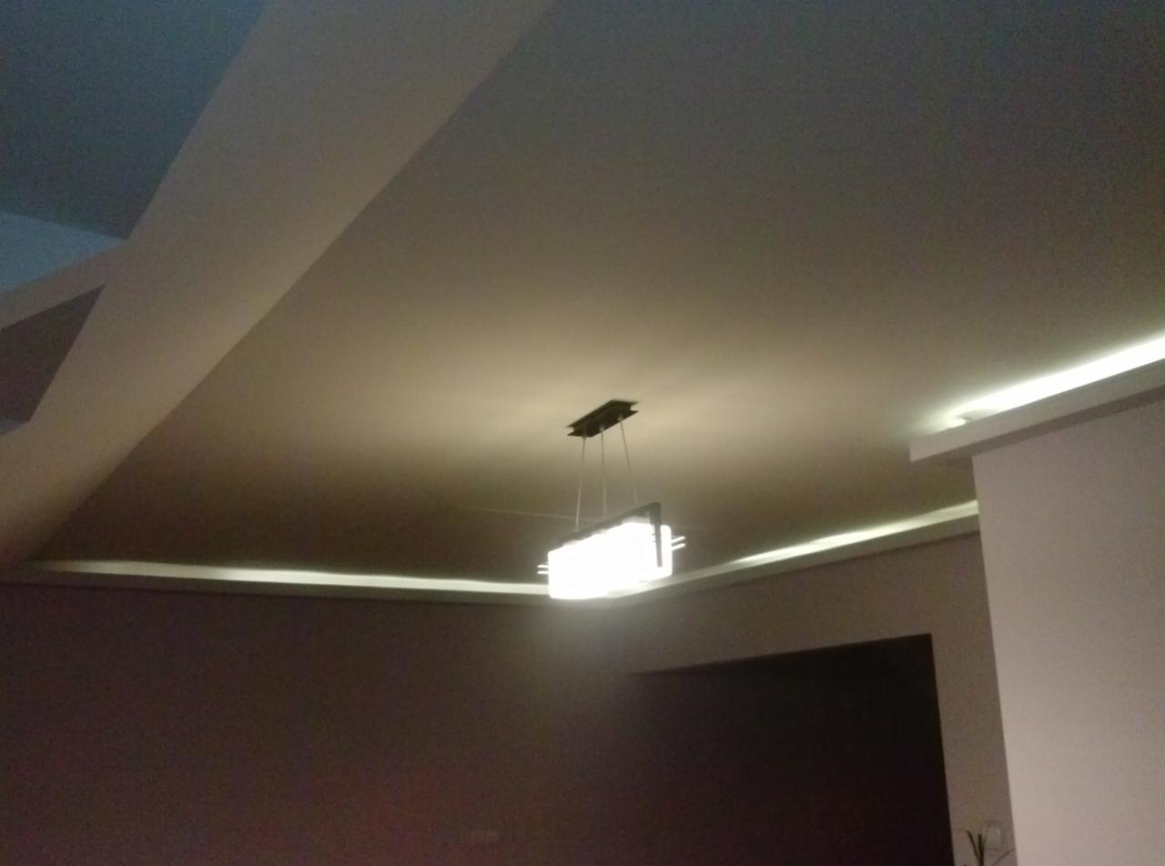 Kremowy sufit podwieszany z oświetleniem LED wzdłuż krawędzi i nowoczesną lampą wiszącą. Nietypowy kształt sufitu z załamaniami.