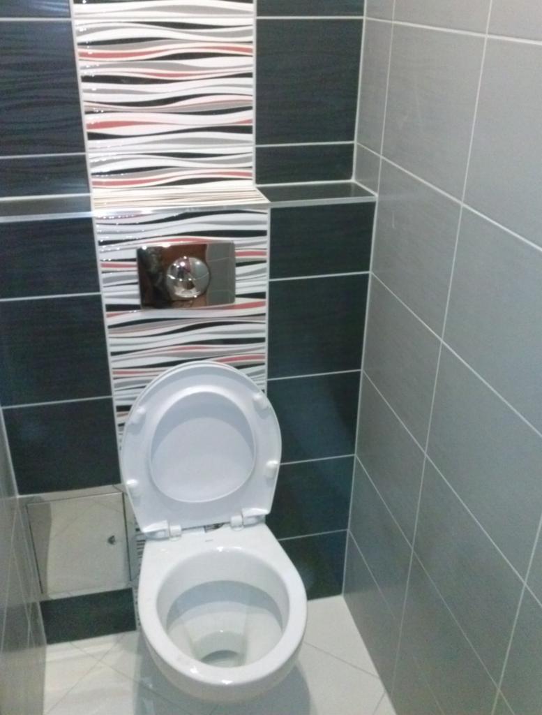 Wykończona łazienka z toaletą i nowoczesnymi płytkami: szare, czarne i białe fale. Widoczny przycisk spłuczki i mała półka. Ukończona usługa glazurnicza.