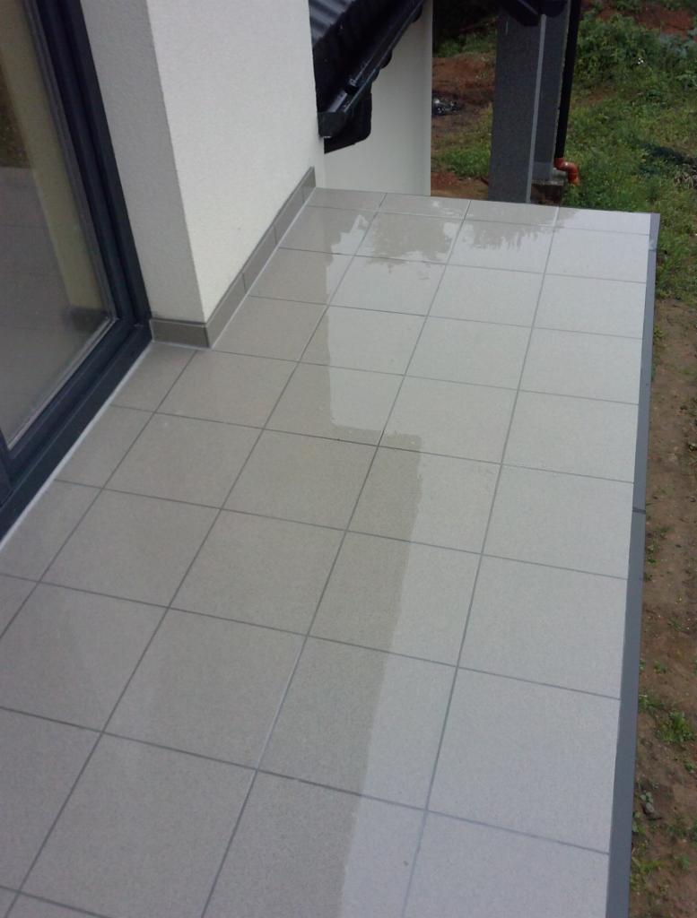 Wyłożony jasnymi płytkami taras przy budynku z szarymi wykończeniami. Widoczne rynna i trawa na zewnątrz. Płytki są mokre, odbijają światło.