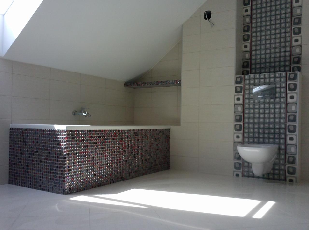 Wykończona łazienka na poddaszu z mozaikową obudową wanny i dekoracyjną płytką na ścianie. Umywalka podwieszana, jasne płytki i skos.