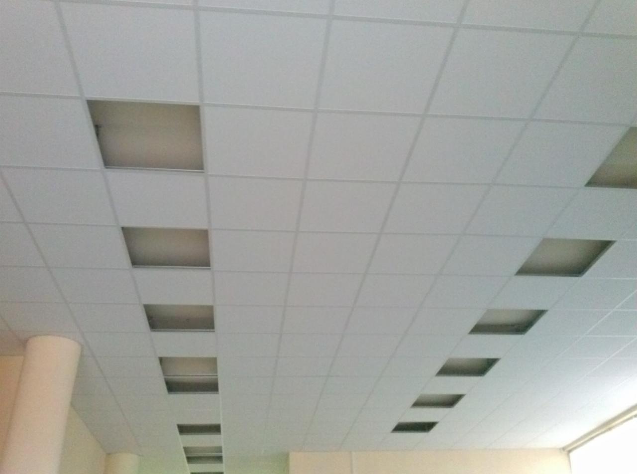Biały sufit podwieszany z kwadratowymi panelami, niektóre z nich zdjęte, ukazując przestrzeń nad sufitem. Widoczny fragment kolumny. Perspektywa z dołu.