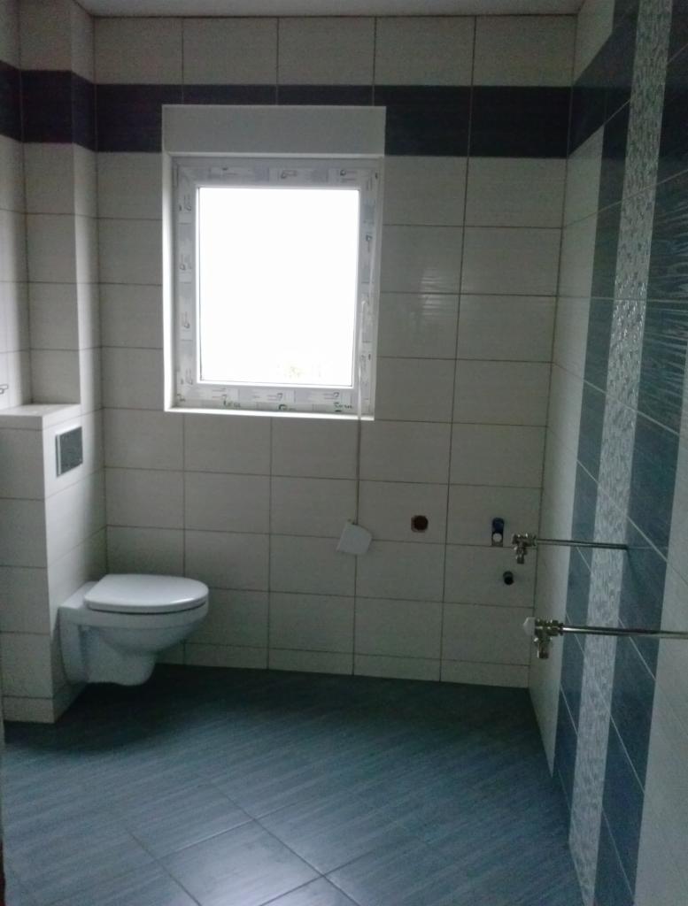 Wykończona łazienka z toaletą podwieszaną, białymi płytkami z ciemnym pasem i szarą podłogą. Widoczne rury i okno z białymi ramami.