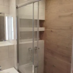 Wykończona łazienka z kabiną prysznicową z przesuwnymi drzwiami, jasnymi płytkami imitującymi marmur i drewnopodobną okładziną ścienną.