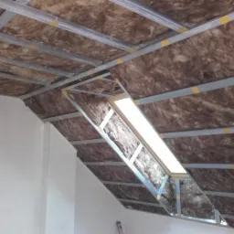 Skośny sufit poddasza z izolacją z wełny mineralnej umieszczoną między metalowymi profilami, widoczny fragment okna dachowego.