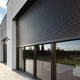 Zewnętrzna roleta aluminiowa w kolorze antracytowym, zasłonięta na oknie tarasowym, widok z zewnątrz na tle jasnej elewacji i błękitnego nieba z chmurami.