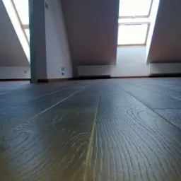 Ciemne panele podłogowe ułożone w pokoju na poddaszu z oknami dachowymi i białymi ścianami. Widoczne grzejniki pod oknami. Wykończenie w kolorze ciemnego drewna.